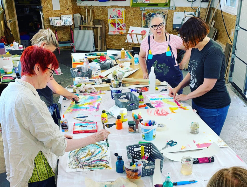 A photograph from an Art Club of several people gathered around a table of supplies. Each is creating their own colourful artwork on a piece of paper.
