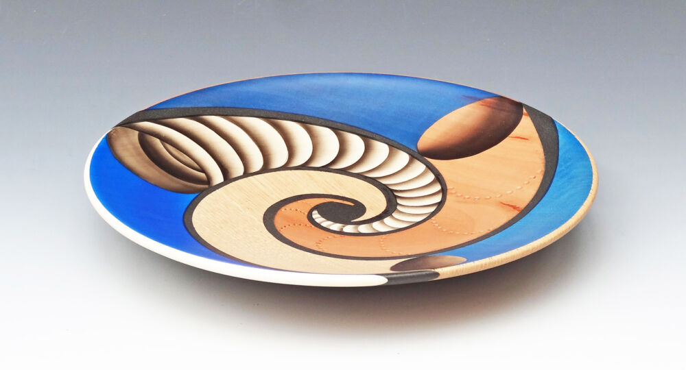 A decorative woodturned plate by Robin Goodman. The design features three horn shaped pieces swirling out from the centre. One piece is reddish brown, one is made from blond wood and the third has dark rings that create a 3D ribbed effect, even though the plate surface is smooth and flat.